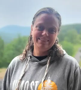 Smiling woman in a gray hoodie with drawstrings outdoors, with green hills in the background used as a backdrop.