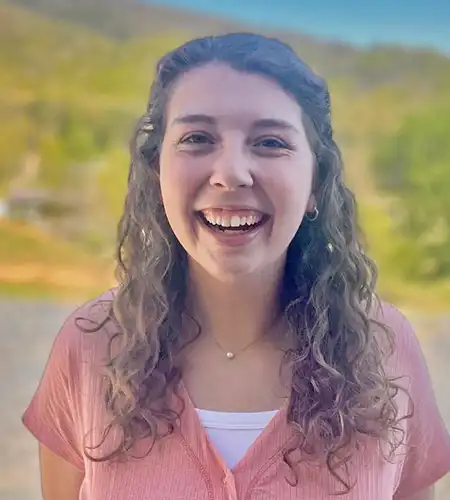 Smiling woman with curly hair in a pink blouse outdoors on a sunny day, background blurred greenery and blue sky.