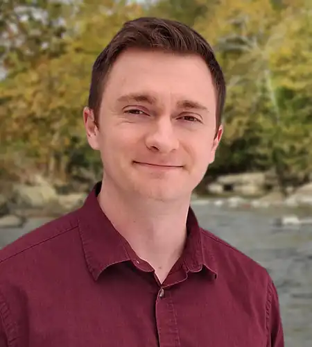 Portrait of a man in a burgundy button-down shirt smiling outdoors by a river with trees in the background