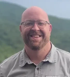 Smiling man with glasses and beard outdoors, grey button-up shirt, mountains blurred in the background